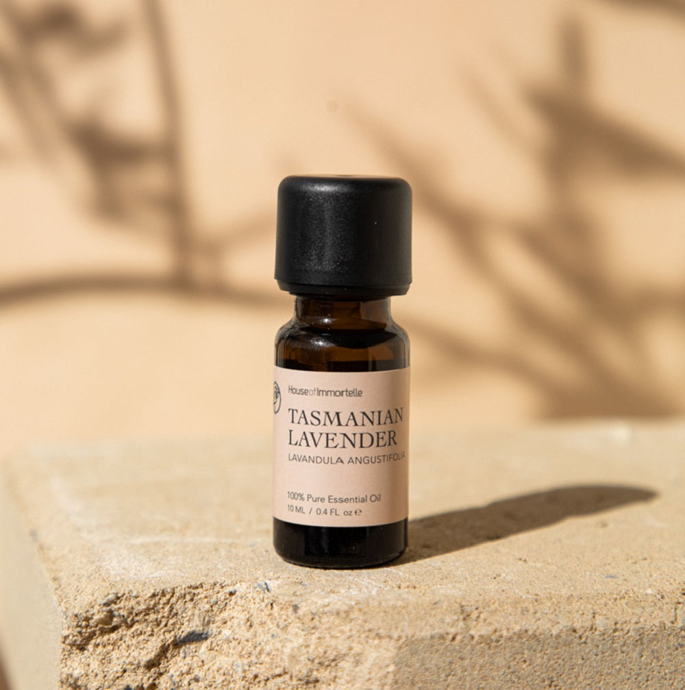 Glass bottle of Tasmanian lavender essential oil with a lavender-colored label, surrounded by sprigs of fresh lavender flowers and soft green leaves, highlighting its calming and soothing properties